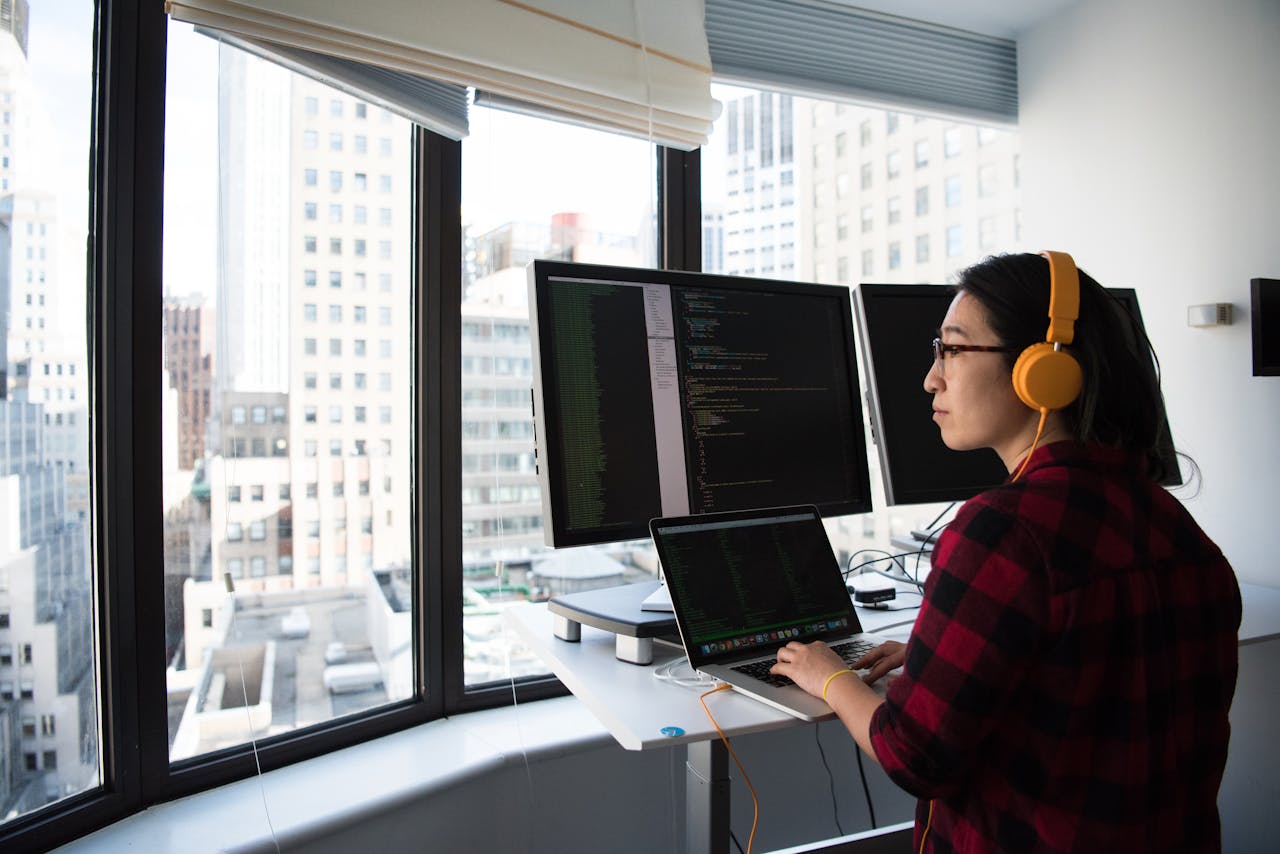 Home A businesswoman coding at a standing desk with city views through large windows, wearing headphones.