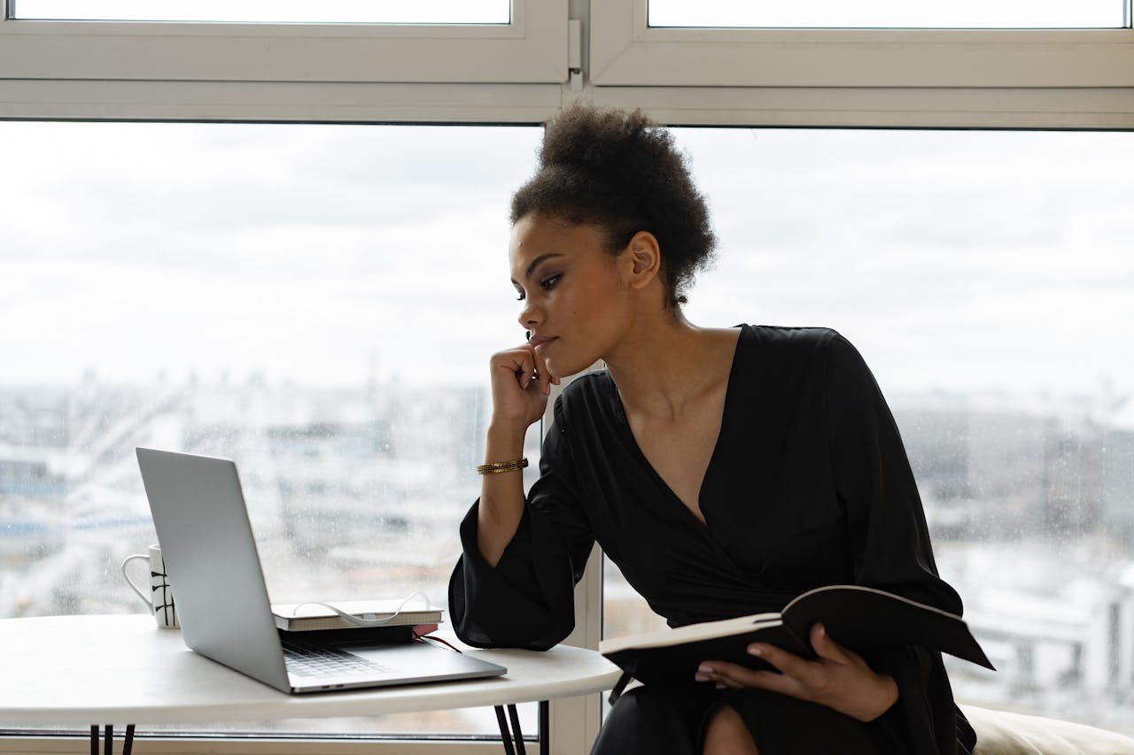 Home A woman in a black dress intently working on a laptop by a large window.